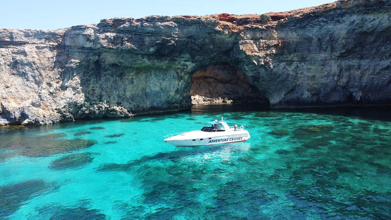 An Adventure Cruises boat anchored in the turquoise water of the Blue Lagoon at Comino, beneath a dramatic natural rock arch.